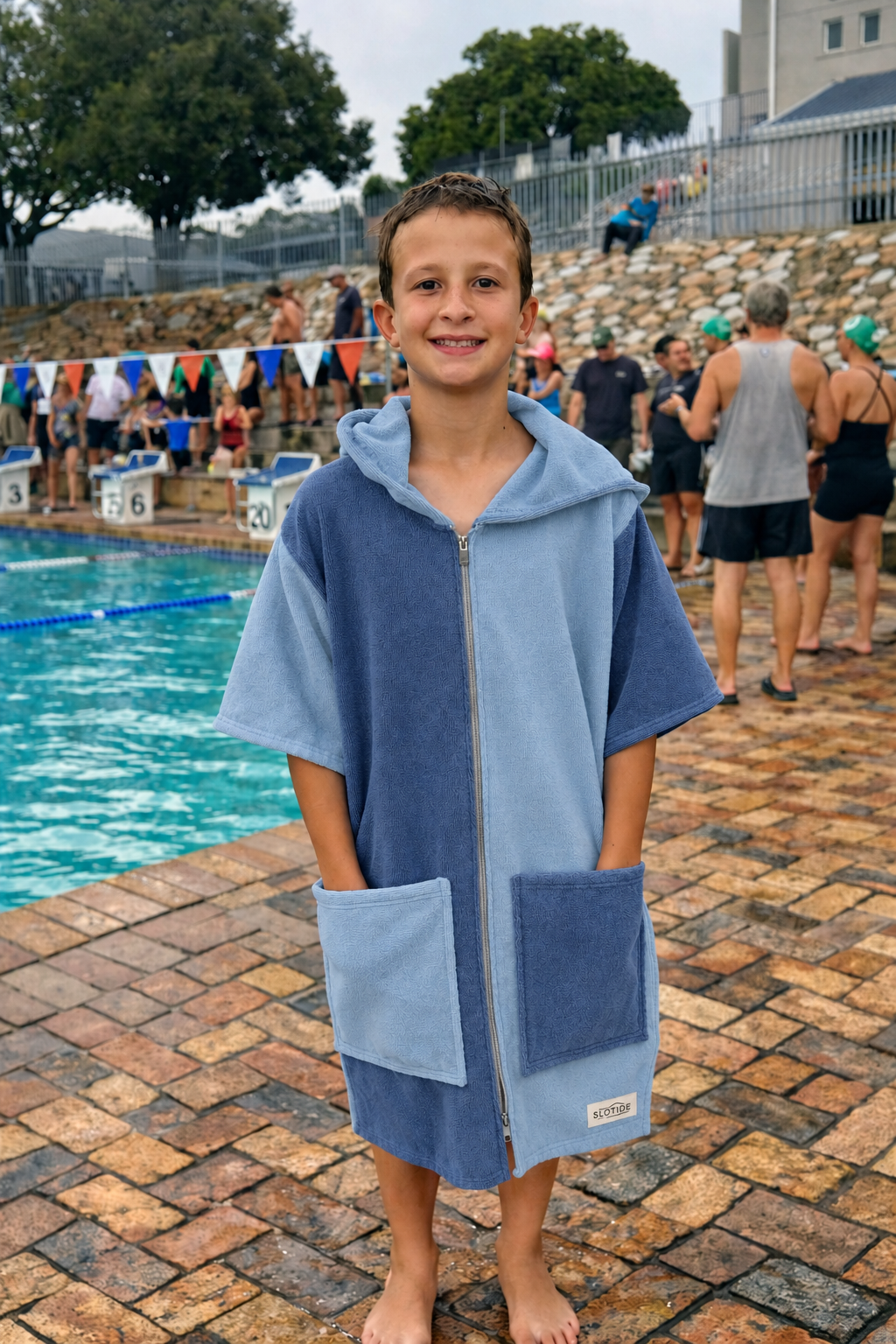 A young boy stands barefoot by an outdoor swimming pool wearing a light blue and medium blue zip-up hooded towel robe with front pockets. Other people and a poolside setting are visible in the background.