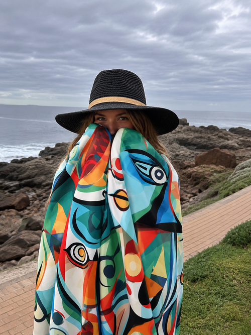 A woman wearing a black wide-brim straw hat and a vibrant, multicolored abstract-patterned wrap or scarf stands on a coastal path with rocks and the ocean in the background.