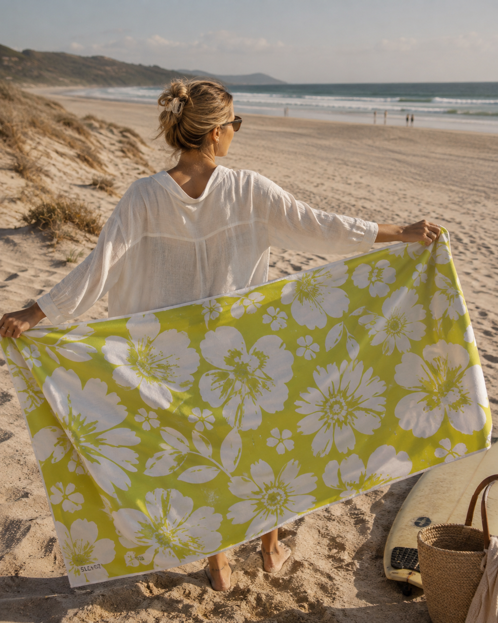 A woman on a sandy beach holds up a large white floral beach towel with a vibrant yellow-green background. She is wearing a white long-sleeve top, with a surfboard and a woven basket bag nearby on the sand.