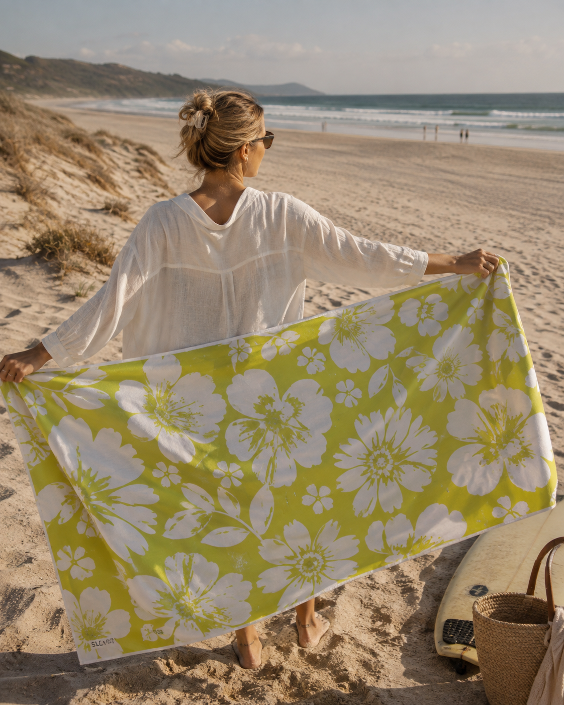 A woman on a sandy beach holds up a large white floral beach towel with a vibrant yellow-green background. She is wearing a white long-sleeve top, with a surfboard and a woven basket bag nearby on the sand.