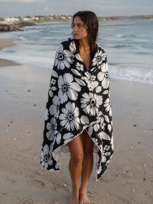 A woman standing on a sandy beach wrapped in a black and white floral print wrap or towel, with the ocean and shoreline in the background.