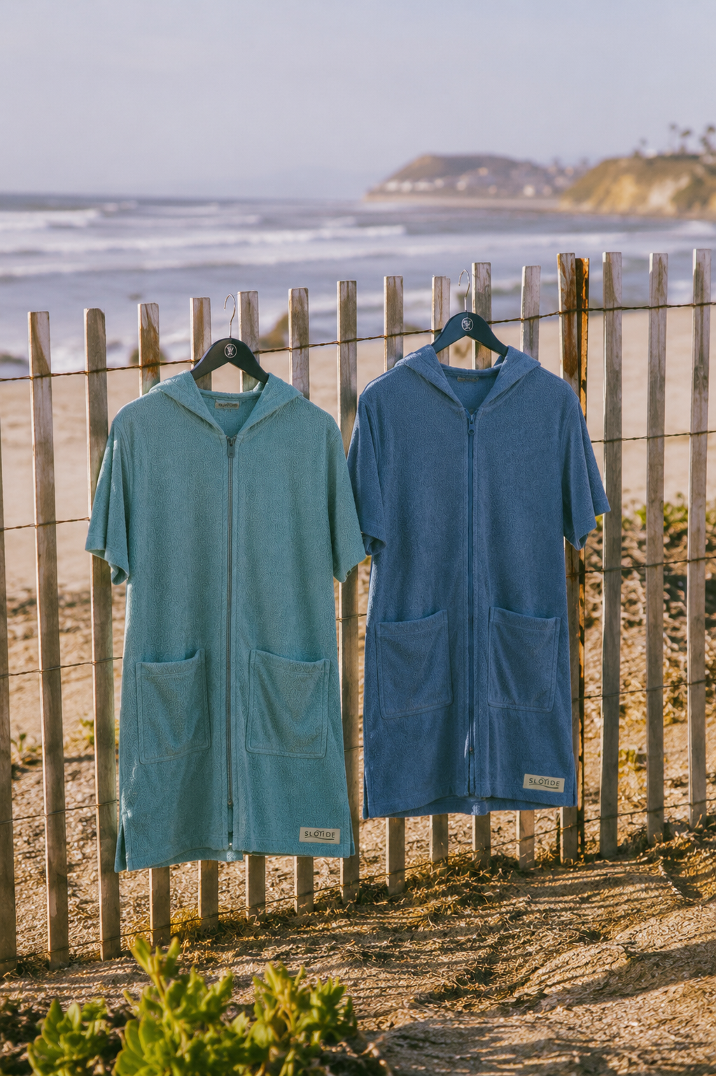 Two hooded zip-front beach towels or robes in teal and medium blue hang on black hangers on a wooden fence at the beach, with ocean waves and a distant cliff visible in the background.