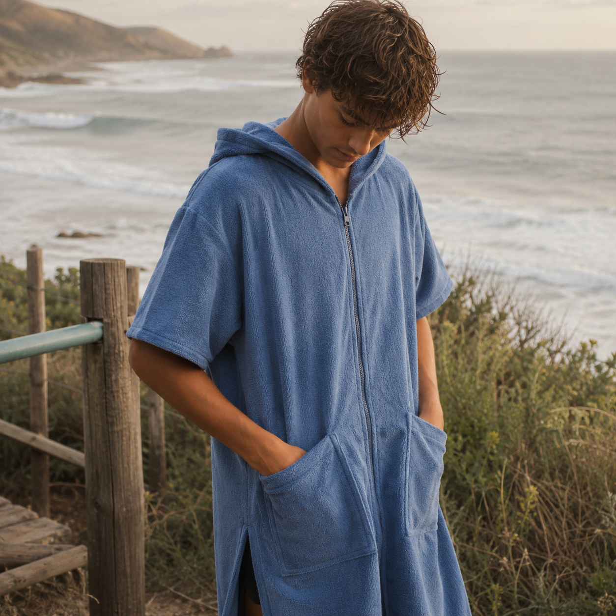 A man wearing a blue zip-up hooded towel robe stands outdoors near the ocean, with dune grass, a wooden fence, and shoreline visible in the background.
