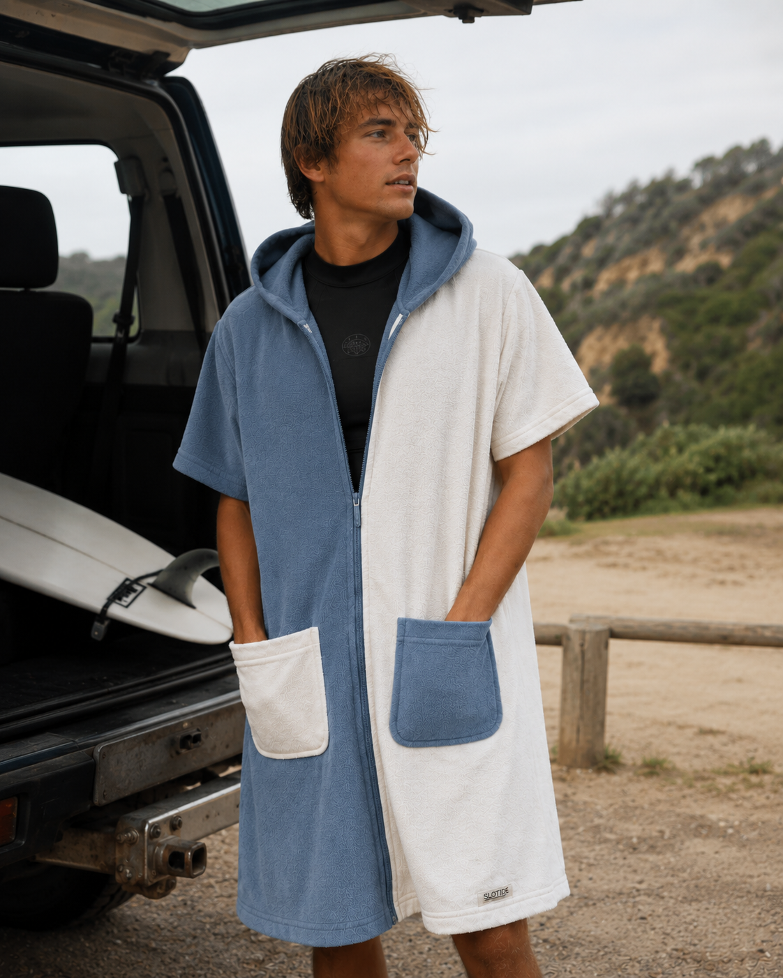A man wearing a cream and blue hooded towel robe stands beside an open vehicle trunk with a surfboard, with a beach and hills in the background. The robe is split vertically with one side in light blue and the other in white, featuring a front zipper, hood, and large patch pockets.