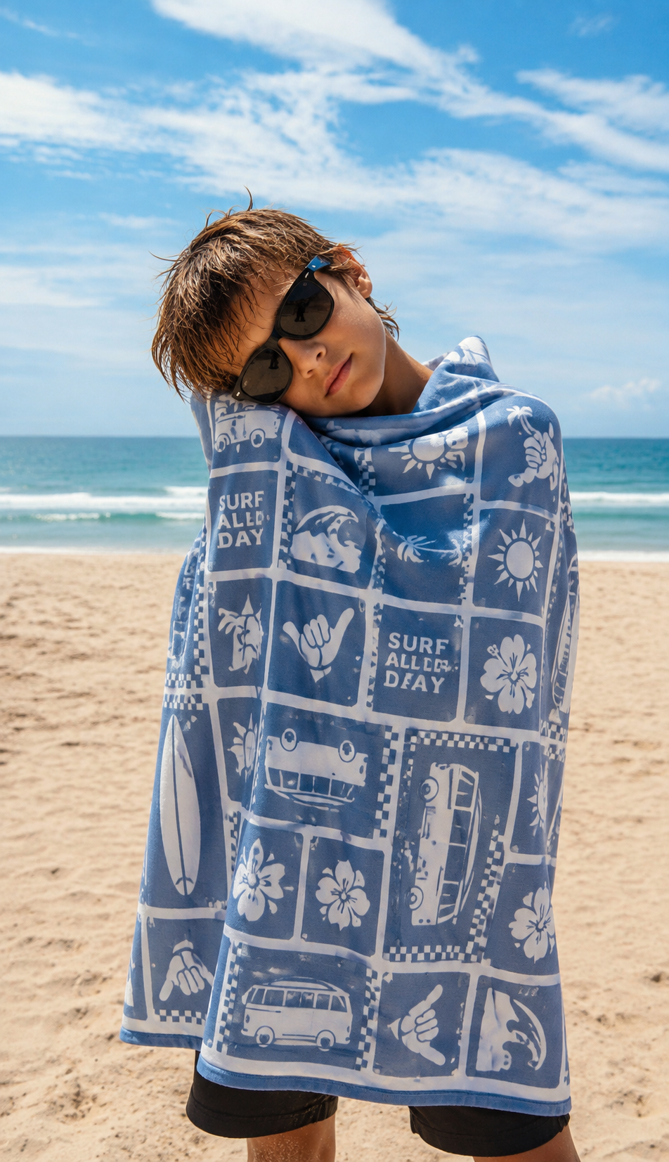 A person standing on a sandy beach wrapped in a blue and white printed towel or wrap featuring surfboard, car, flower, and 'SURF ALL DAY' graphics, with ocean and blue sky in the background.
